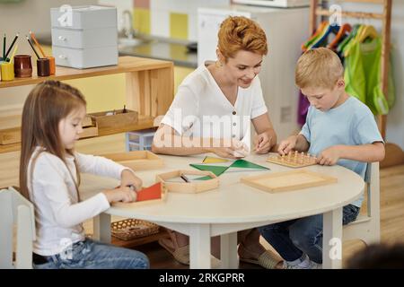 smiling teacher playing with kids and didactic materials on table in montessori school Stock Photo