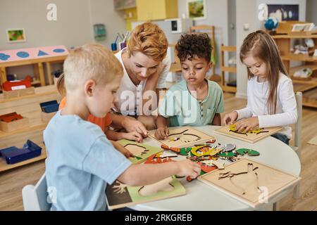 teacher talking to interracial children playing together near game on table in montessori school Stock Photo