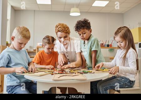 smiling teacher playing with multiethnic kids near game on table in montessori school Stock Photo