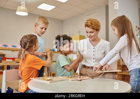 smiling multiethnic kids holding didactic materials near teacher in montessori school Stock Photo