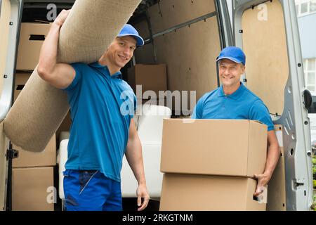 Two Male Workers Carrying Carpet And Cardboard Boxes In Front Of Van Stock Photo
