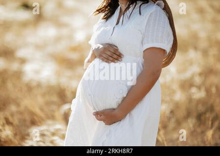 A closeup of a pregnant woman in a white dress, posing in a wheat field - motherhood concept Stock Photo