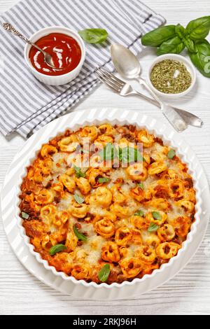 tortellini bake with mozzarella cheese, tomato sauce and ground beef in white baking dish on white wooden table with ketchup, vertical view from above Stock Photo