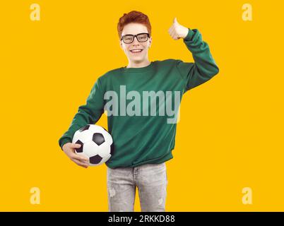 Smiling, happy red-haired boy holds soccer ball on isolated yellow background. Stock Photo