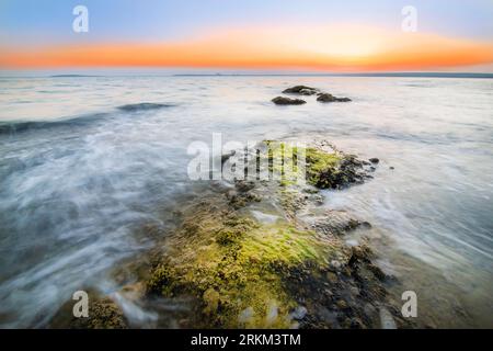 A beautiful sunset at sea with mossy rocks in the reflective water ...