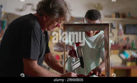 Young carpenter working at workshop with the help of his teacher. Senior man guiding pupil with furniture building, woodworking job occupation Stock Photo