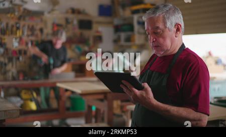 Senior worker wearing apron standing inside carpenter workshop holding tablet and browsing orders, job occupation concept using modern technology Stock Photo