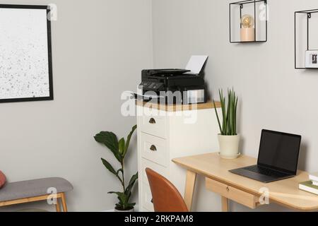 Modern printer with paper on chest of drawers near wooden table indoors Stock Photo