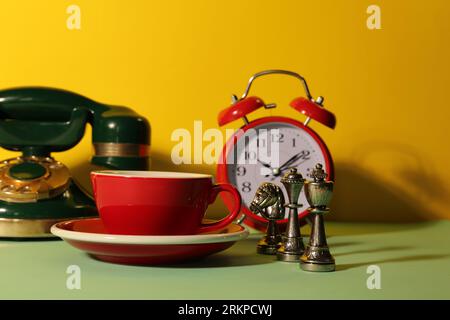 Red cup with saucer, alarm clock, vintage telephone and chessmen on light green table against yellow background Stock Photo