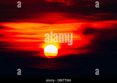 Intense bright sun setting through heavy dark clouds creating a fiery red and orange sky background texture silhouette Stock Photo