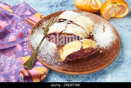 Homemade festive cherry strudel pastry with powdered sugar on rustic brown plate, with tangerines, green tea leaves, vintage silver fork, on the blue Stock Photo