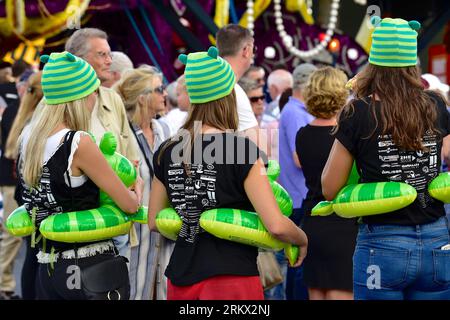 Three girls with green striped hats and floaties around their waists are standing among the audience watching demonstrations of Flower Parade float Stock Photo