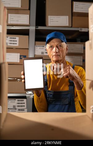 Old inventory warehouse manager holding digital tablet mockup blank white screen pointing finger. Over boxes Stock Photo