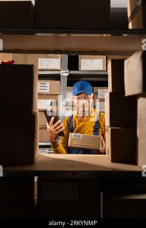 Elderly warehouse worker inventory manager using digital tablet works standing near shelves full of parcels ready to be shipped Stock Photo