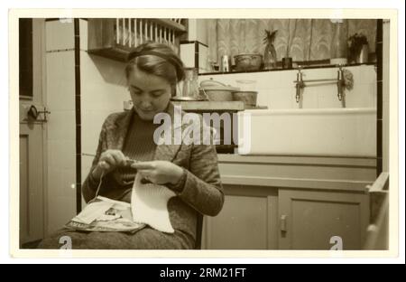 Original 1950's era photograph of a young woman sitting in her kitchen, wearing a tweed suit and skinny ribbed jumper underneath. A working woman or possibly in student digs. The young lady is knitting, using a wool pattern, U.K. Stock Photo