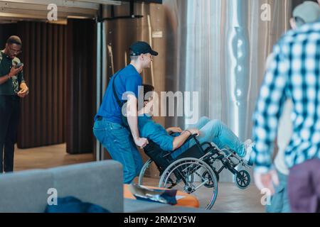Side photo of positive male employees having fun at work during break. Young male person wearing cap pushing his male coworker in wheelchair at the of Stock Photo