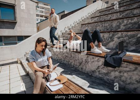 Tired businessman lying on a bench and reading paperwork outdoors. Brunette woman working on tablet remotely. Blonde businessman having phone call whi Stock Photo