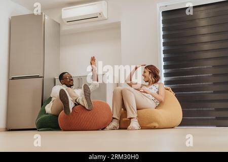 Happy, tired, diverse colleagues taking a break from work at the office, lying on lazy bags, having a conversation, giving high five each other. Stock Photo