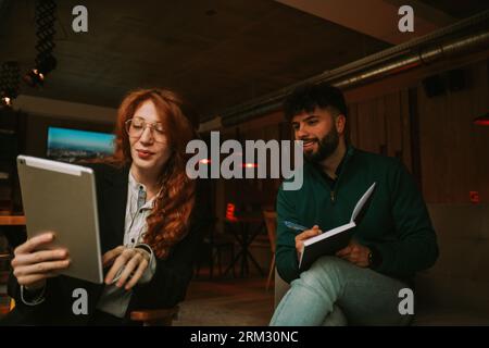 Beautiful smiley ginger female person showing something on the lap top to her male colleague while working together at the coffee bar on their new pro Stock Photo
