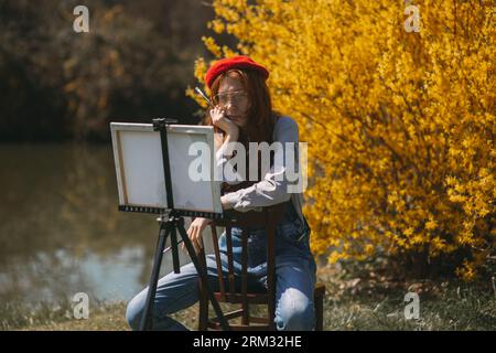Beautiful ginger girl with red beret sitting on a wooden chair with her artwork displayed on an easel, thinking and looking at it Stock Photo