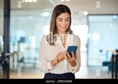Smiling mid aged business woman executive using smartphone standing in office. Stock Photo