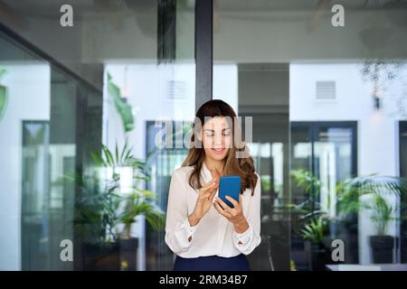 Beautiful mature business woman executive using smartphone walking in office. Stock Photo