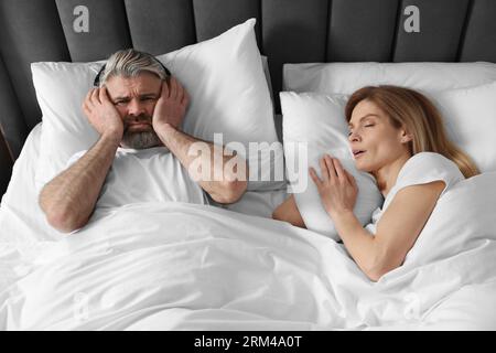 Irritated man with headphones lying near his snoring wife in bed at home Stock Photo