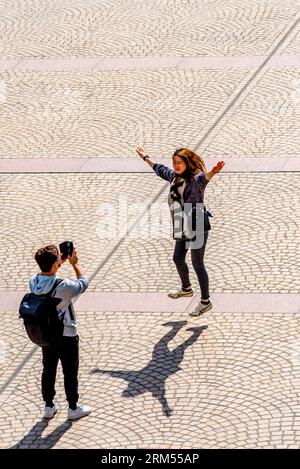 A young man uses a mobile phone to photograph a young woman doing a star jump on the forecourt area of the Sydney Opera House in Australia Stock Photo
