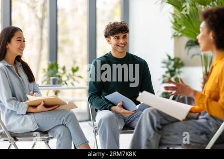 Happy multiracial students friends talking and discussing educational project together, sitting on chairs in circle Stock Photo