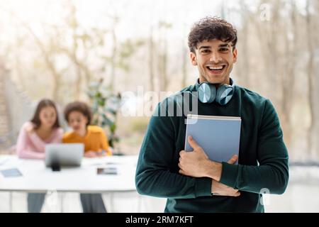 Portrait of cheerful male university student posing with notepads and smiling while groupmates studying on background Stock Photo
