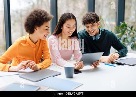 Happy three international students sitting at desk in audience and using digital tablet, discussing project together Stock Photo