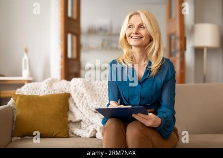 Blonde mature woman holding clipboard and taking notes indoor Stock Photo