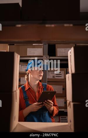 Manager checking inventory uses digital tablet. Young woman working in warehouse with rows of shelves full of parcels. Stock Photo