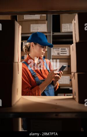 Female warehouse worker using mobile app checking parcel box. Entrepreneur seller holding cell phone tech device while standing near shelves with Stock Photo