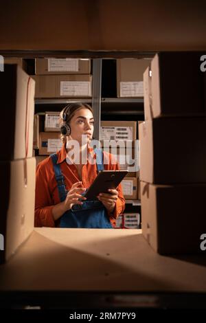 Young female worker wearing headset in logistics distribution warehouse using digital tablet over boxes background Stock Photo