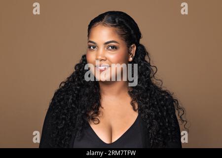 Headshot portrait of young body positive brazilian woman with wavy hair smiling at camera, standing on brown background Stock Photo