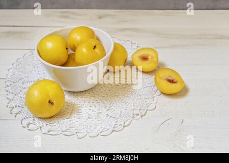A white porcelain bowl filled with ripe yellow plums on a crocheted cloth on a white board Stock Photo