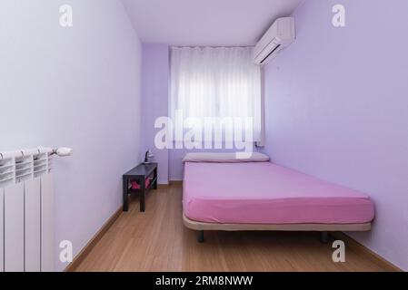 Bedroom with a single bed without clothes, an air conditioner, a window with curtains, wooden floors and an aluminum radiator Stock Photo