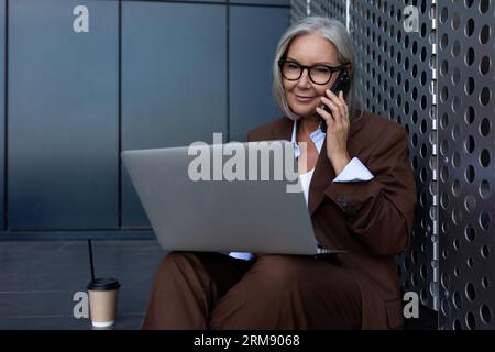 mature business woman with gray hair dressed in a brown suit is holding an online meeting using a laptop in the street Stock Photo