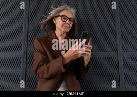 confident charming mature old lady with gray hair and glasses is dressed in a stylish jacket in an urban environment Stock Photo