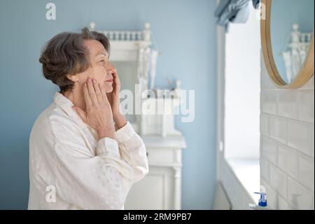 Elderly woman applying anti-age cream on face in bathroom Stock Photo