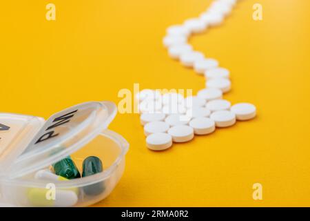 Arrow shaped white medical pills point to the plastic daily organizer (for a week of medication) and flow towards it. Close-up on a yellow background. Stock Photo