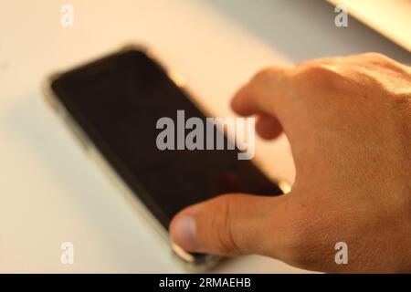 A photo of a hand reaching towards a smartphone on a white desk. Stock Photo