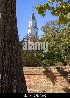 Bruton Parish Church's spire rises in colonial Williamsburg, Virginia, embraced by a blue sky and encircled by a tree trunk and lush leaves. Stock Photo