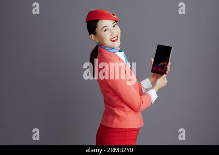happy stylish asian female stewardess in red skirt, jacket and hat uniform using tablet PC against gray background. Stock Photo