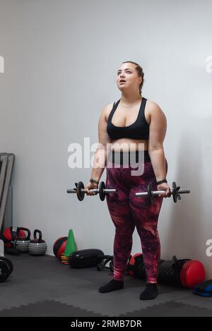 A focused young woman engages in strength training at a gym. Holding weights in both hands, her determination and concentration are evident as she str Stock Photo