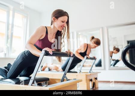 Two young females doing ABS muscles strength exercises using  pilates reformer machine in sport athletic gym hall. Active people training, yoga classe Stock Photo