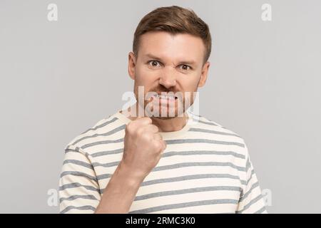 Angry aggressive middle aged man clenched fist has evil face looking at camera on grey background. Stock Photo