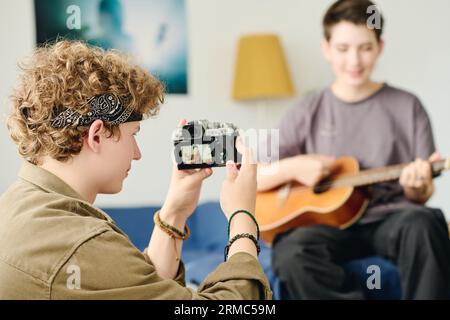 Focus on teenage guy with camera taking photo or shooting video of his girlfriend playing acoustic guitar at home while sitting in front of her Stock Photo
