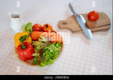 A plate of fresh vegetables, a knife, and a wooden chopping board on a dining table. Sweet peppers, carrots, tomatoes, and green lettuce. Healthy home Stock Photo